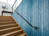 Papier peint bois bleu vieilli posé le long d'un escalier aux marches en chêne avec main courante noire
